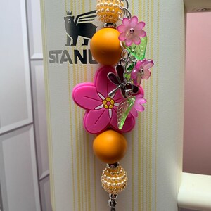 May include: A decorative tumbler charm featuring a large pink flower bead, orange spherical beads, and textured cream beads. The charm includes small translucent pink flower charms, green leaf charms, and a silver dragonfly charm on a metal chain.