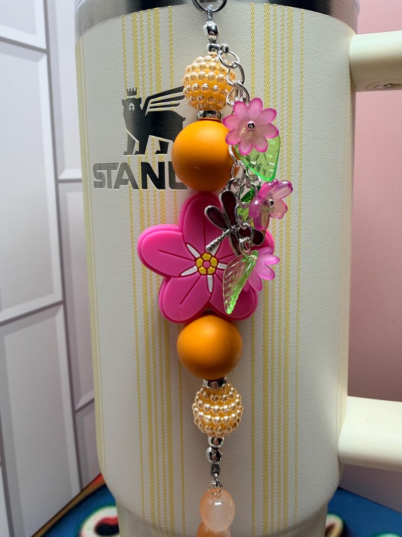 May include: A decorative tumbler charm featuring a large pink flower bead, orange spherical beads, and textured cream beads. The charm includes small translucent pink flower charms, green leaf charms, and a silver dragonfly charm on a metal chain.