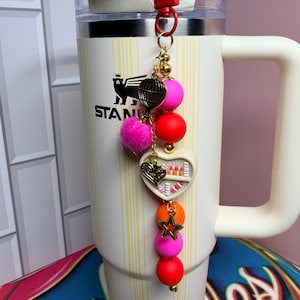 K&ouml;nnte beinhalten: Ein dekorativer Schl&uuml;sselanh&auml;nger mit rotem Verschluss und goldener Kette. Der Anh&auml;nger hat einen rosa Pompon, einen herzf&ouml;rmigen Anh&auml;nger und bunte Perlen in Pink, Orange und Rot. Der Schl&uuml;sselanh&auml;nger ist an einem wei&szlig;en Becher mit Griff befestigt.
