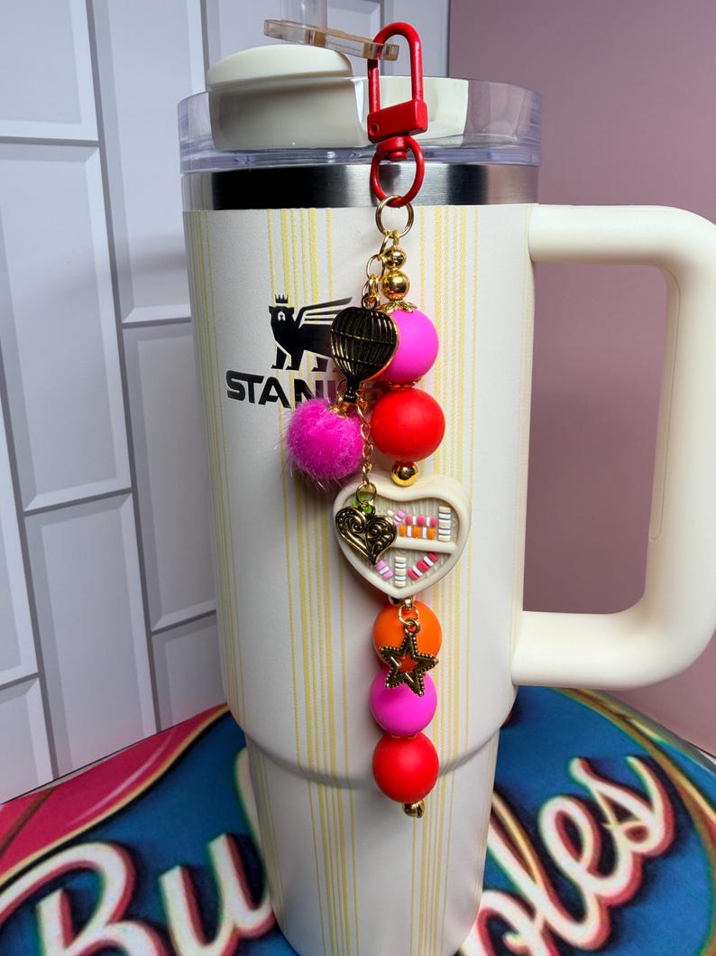 K&ouml;nnte beinhalten: Ein dekorativer Schl&uuml;sselanh&auml;nger mit rotem Verschluss und goldener Kette. Der Anh&auml;nger hat einen rosa Pompon, einen herzf&ouml;rmigen Anh&auml;nger und bunte Perlen in Pink, Orange und Rot. Der Schl&uuml;sselanh&auml;nger ist an einem wei&szlig;en Becher mit Griff befestigt.