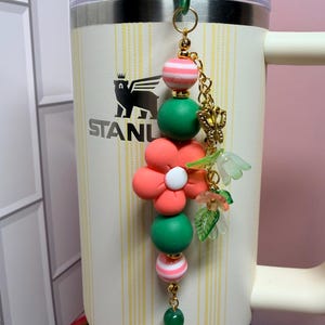May include: A decorative tumbler charm featuring a vertical arrangement of round beads in pink and white stripes and solid green, centered by a large coral-colored flower bead with a white center, accented by a gold butterfly and small green leaf charms.