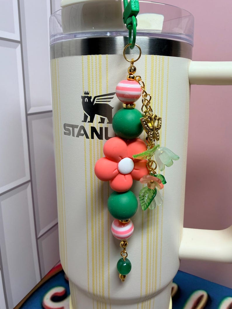 May include: A decorative tumbler charm featuring a vertical arrangement of round beads in pink and white stripes and solid green, centered by a large coral-colored flower bead with a white center, accented by a gold butterfly and small green leaf charms.