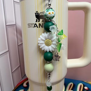 May include: A cream-colored Stanley tumbler with vertical yellow stripes features a decorative beaded charm attached to the lid. The charm includes a large white daisy bead, green silicone spheres, floral-shaped beads, and a small silver butterfly charm.