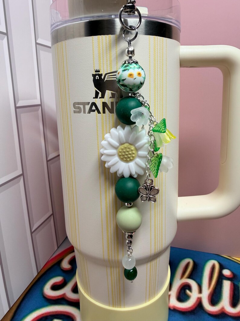 May include: A cream-colored Stanley tumbler with vertical yellow stripes features a decorative beaded charm attached to the lid. The charm includes a large white daisy bead, green silicone spheres, floral-shaped beads, and a small silver butterfly charm.