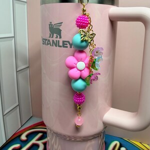 May include: A light pink Stanley tumbler with a handle and a decorative beaded charm attached to the lid. The charm features a large pink flower bead, turquoise round beads, textured hot pink beads, a gold butterfly charm, and small floral accents.