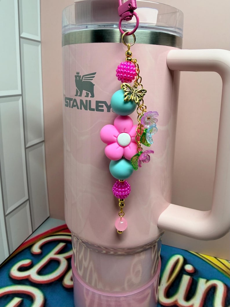May include: A light pink Stanley tumbler with a handle and a decorative beaded charm attached to the lid. The charm features a large pink flower bead, turquoise round beads, textured hot pink beads, a gold butterfly charm, and small floral accents.