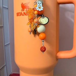 May include: An orange tumbler with a handle and a lid, featuring a decorative charm attached to the top. The charm includes a white ghost, a green pumpkin, orange maple leaves, and various orange, white, and glittery beads on a silver chain.