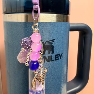 May include: A decorative charm featuring a small clear vial filled with purple glitter, accented with a silver turtle charm, a purple bead, and a strand of pink and purple gradient beads. The charm is attached to a light purple plastic lobster clasp.