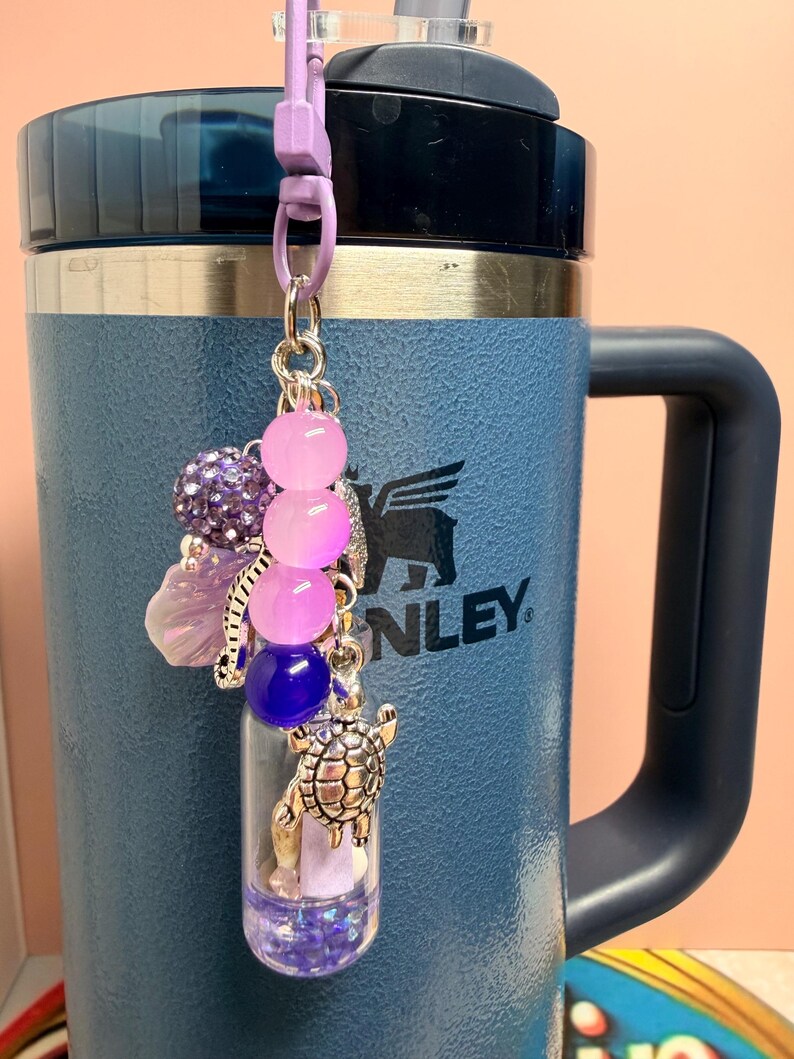 May include: A decorative charm featuring a small clear vial filled with purple glitter, accented with a silver turtle charm, a purple bead, and a strand of pink and purple gradient beads. The charm is attached to a light purple plastic lobster clasp.