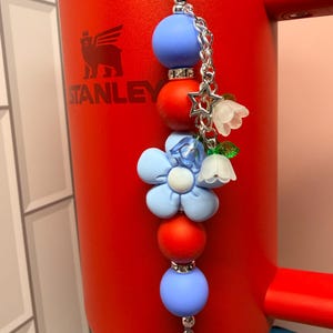 Op de afbeelding: Een decoratieve sleutelhanger met een ketting en diverse bedels. De bedels omvatten blauwe en rode kralen, een lichtblauwe bloem, een witte bloem en een zilveren ster. De sleutelhanger hangt aan een rode Stanley-beker.