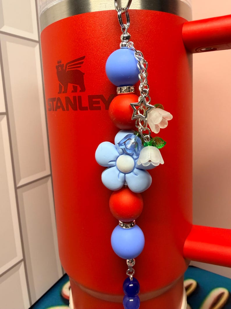 Op de afbeelding: Een decoratieve sleutelhanger met een ketting en diverse bedels. De bedels omvatten blauwe en rode kralen, een lichtblauwe bloem, een witte bloem en een zilveren ster. De sleutelhanger hangt aan een rode Stanley-beker.