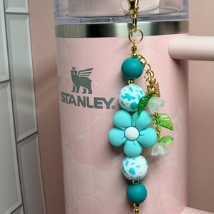May include: A decorative keychain featuring teal and white speckled silicone beads, a large teal flower-shaped bead, and small translucent green leaf charms. The accessory is attached to a gold-toned metal lobster clasp and hangs against a light pink Stanley tumbler.
