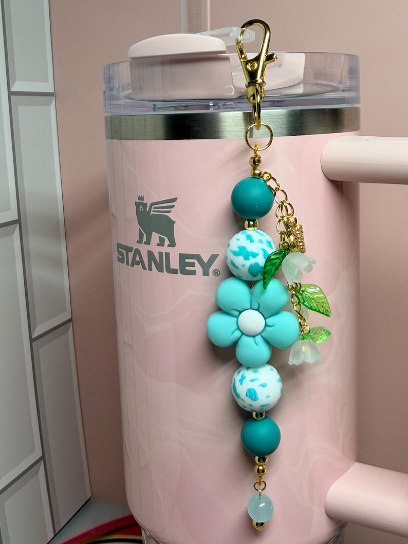 May include: A decorative keychain featuring teal and white speckled silicone beads, a large teal flower-shaped bead, and small translucent green leaf charms. The accessory is attached to a gold-toned metal lobster clasp and hangs against a light pink Stanley tumbler.