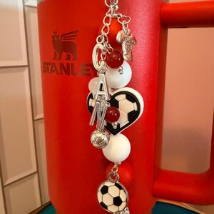 Puede incluir: Un dije decorativo para vaso con una cadena plateada, cuentas rojas y blancas, un dije de bal&oacute;n de f&uacute;tbol, un dije de bal&oacute;n en forma de coraz&oacute;n, un dije de camiseta plateada y letras plateadas que forman CAN, colgado de un vaso rojo Stanley.