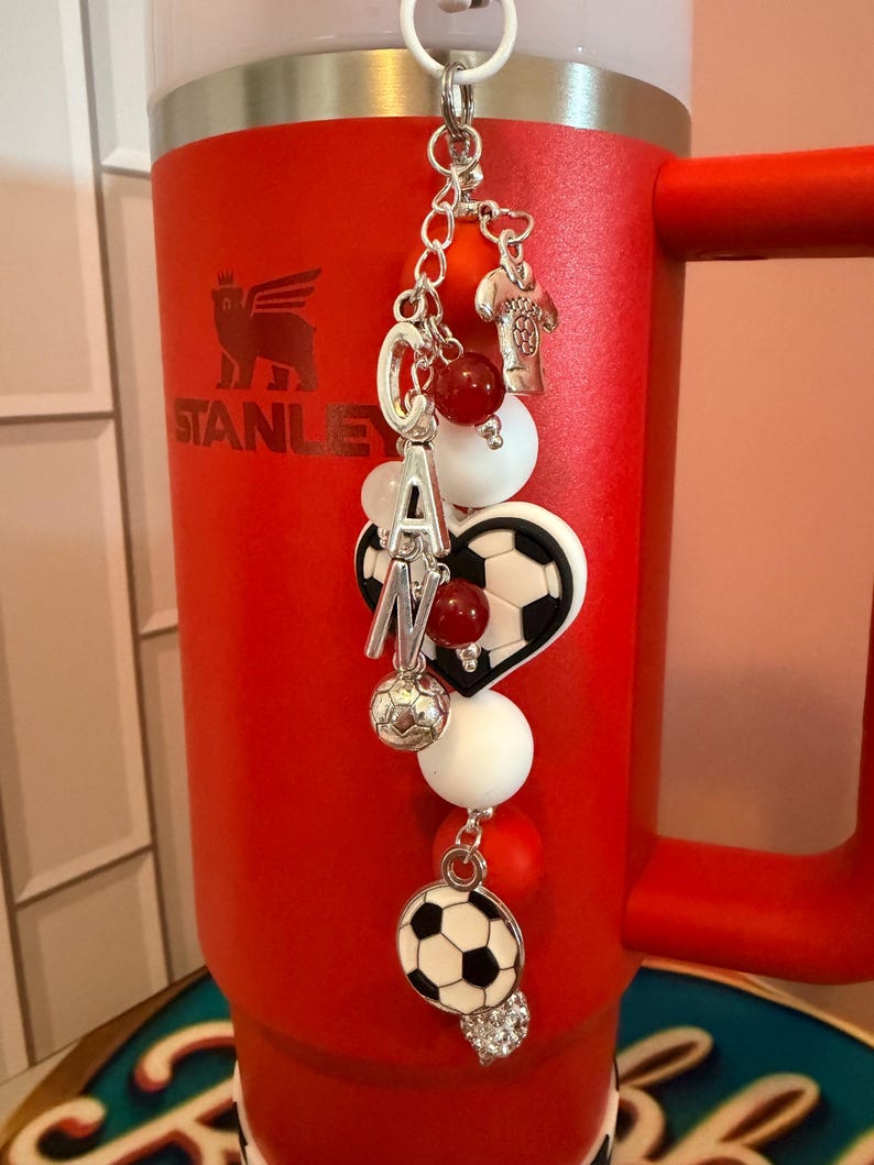 Puede incluir: Un dije decorativo para vaso con una cadena plateada, cuentas rojas y blancas, un dije de bal&oacute;n de f&uacute;tbol, un dije de bal&oacute;n en forma de coraz&oacute;n, un dije de camiseta plateada y letras plateadas que forman CAN, colgado de un vaso rojo Stanley.