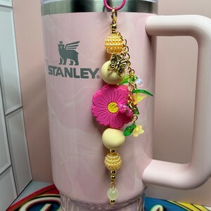 May include: A light pink Stanley tumbler with a handle and a clear lid, featuring a decorative charm attached to the top. The charm consists of a gold-tone chain with yellow textured beads, a pink flower, and small green leaf accents.