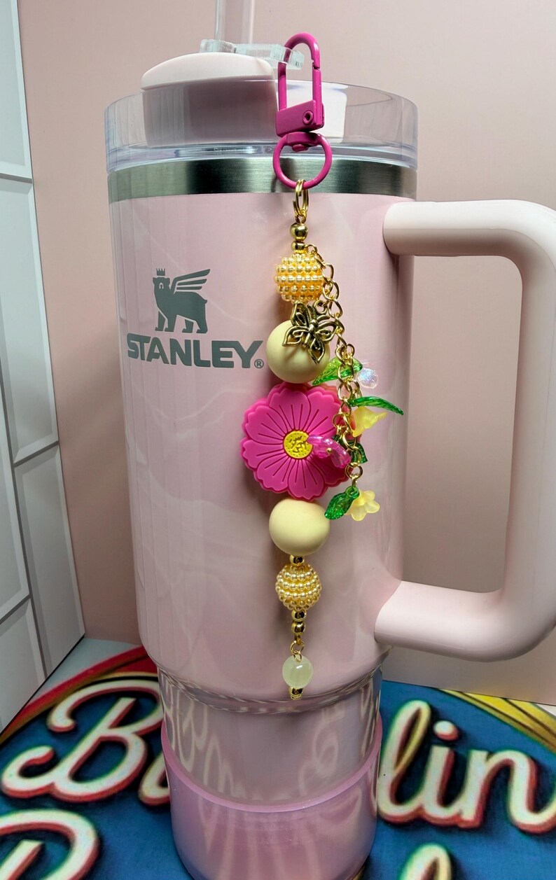 May include: A light pink Stanley tumbler with a handle and a clear lid, featuring a decorative charm attached to the top. The charm consists of a gold-tone chain with yellow textured beads, a pink flower, and small green leaf accents.