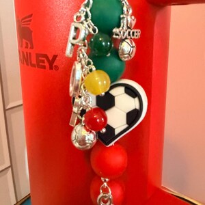 Puede incluir: Un accesorio decorativo con una cadena plateada, cuentas verdes, amarillas y rojas, y dijes plateados que incluyen un bal&oacute;n de f&uacute;tbol, la letra P y un dije en forma de coraz&oacute;n con un patr&oacute;n de bal&oacute;n de f&uacute;tbol en blanco y negro.