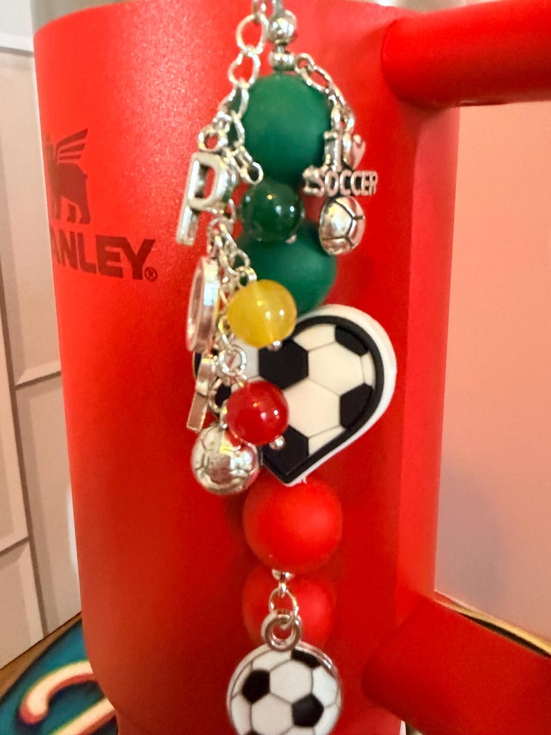 Puede incluir: Un accesorio decorativo con una cadena plateada, cuentas verdes, amarillas y rojas, y dijes plateados que incluyen un bal&oacute;n de f&uacute;tbol, la letra P y un dije en forma de coraz&oacute;n con un patr&oacute;n de bal&oacute;n de f&uacute;tbol en blanco y negro.