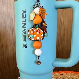 May include: A light blue Stanley tumbler featuring a vertical charm accessory with orange and white beads, a pumpkin-shaped pendant, and a translucent orange leaf charm. A beaded band with a floral and pumpkin pattern encircles the base of the cup.