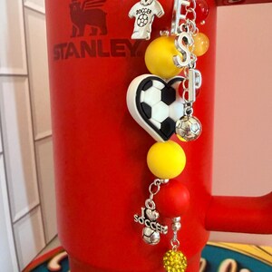 Puede incluir: Un vaso Stanley de color rojo brillante con un accesorio de dijes con tem&aacute;tica de f&uacute;tbol. El dije incluye cuentas amarillas y rojas, letras plateadas que forman ESP, un bal&oacute;n en forma de coraz&oacute;n, un dije de camiseta y uno de I Love Soccer.