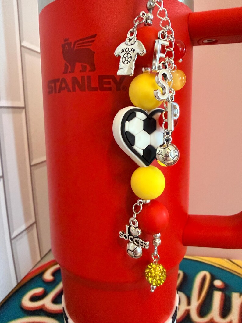Puede incluir: Un vaso Stanley de color rojo brillante con un accesorio de dijes con tem&aacute;tica de f&uacute;tbol. El dije incluye cuentas amarillas y rojas, letras plateadas que forman ESP, un bal&oacute;n en forma de coraz&oacute;n, un dije de camiseta y uno de I Love Soccer.