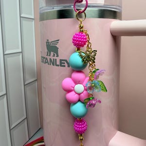 May include: A decorative keychain featuring a pink and turquoise bead strand with a central pink flower charm, a gold-toned butterfly charm, and small translucent purple and blue flower accents, all attached to a pink metal lobster clasp.