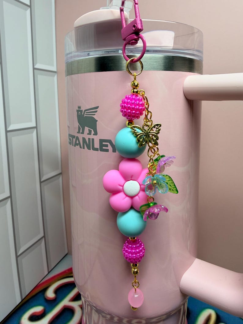 May include: A decorative keychain featuring a pink and turquoise bead strand with a central pink flower charm, a gold-toned butterfly charm, and small translucent purple and blue flower accents, all attached to a pink metal lobster clasp.