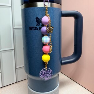 May include: A navy blue insulated tumbler with a handle and a decorative charm. The charm features a series of colourful beads, a gold butterfly, and a purple acrylic tag that says "Hello Spring".