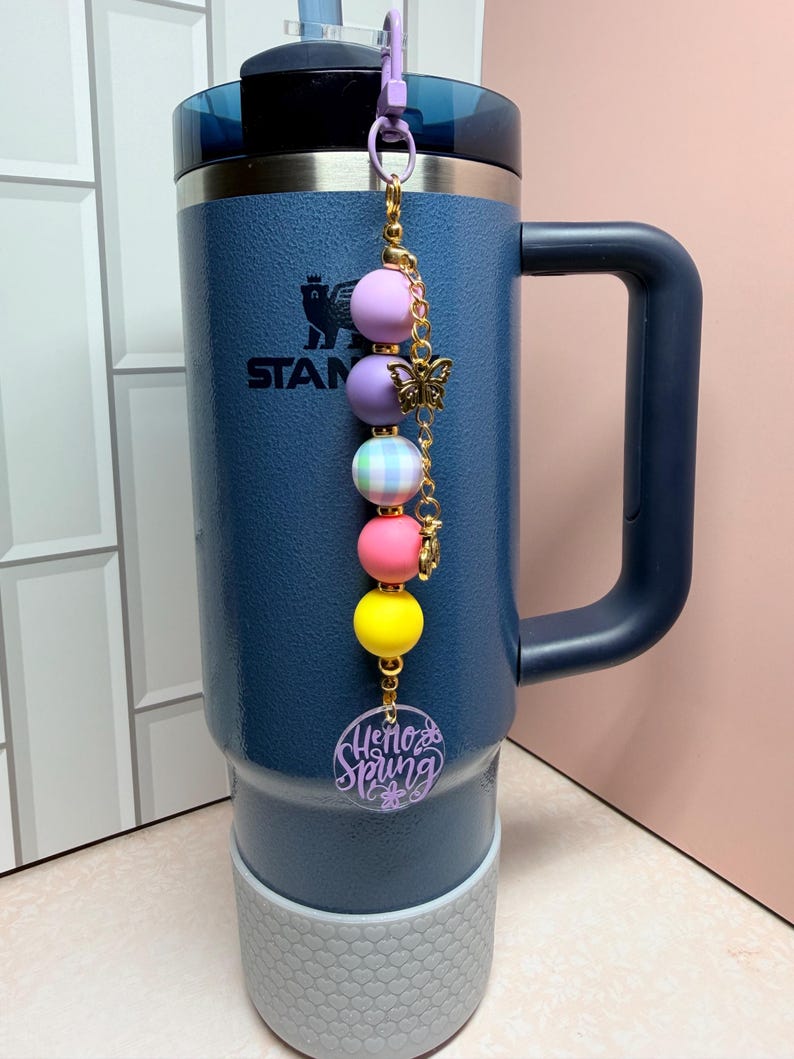 May include: A navy blue insulated tumbler with a handle and a decorative charm. The charm features a series of colourful beads, a gold butterfly, and a purple acrylic tag that says "Hello Spring".