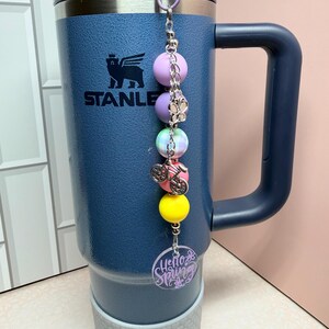 May include: A navy blue Stanley tumbler with a decorative charm. The charm features a series of colourful beads, a bicycle charm, and a purple circular tag that reads "Hello Spring". The tumbler has a grey base and a black handle.