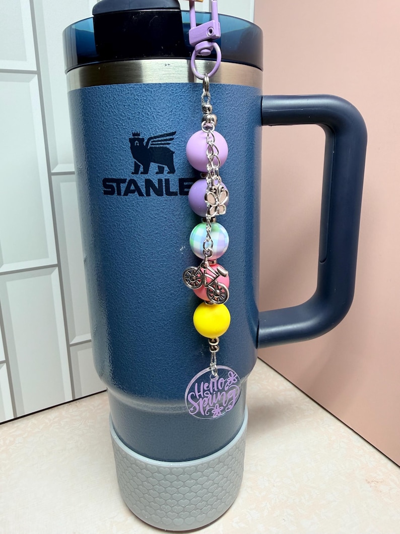 May include: A navy blue Stanley tumbler with a decorative charm. The charm features a series of colourful beads, a bicycle charm, and a purple circular tag that reads "Hello Spring". The tumbler has a grey base and a black handle.