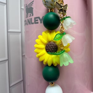 May include: A decorative charm featuring a vertical arrangement of beads including a floral patterned sphere, dark green spheres, a large yellow sunflower, and a white sphere. A gold-toned metal butterfly and three translucent bell-shaped flowers hang from the side.