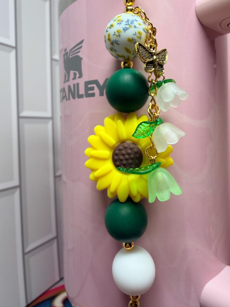 May include: A decorative charm featuring a vertical arrangement of beads including a floral patterned sphere, dark green spheres, a large yellow sunflower, and a white sphere. A gold-toned metal butterfly and three translucent bell-shaped flowers hang from the side.