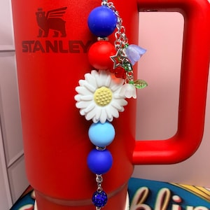 Op de afbeelding: Een rode Stanley-beker met een decoratieve bedel. De bedel heeft blauwe, rode en witte kralen, een witte madelief en kleine bloemaccenten. De beker heeft een handvat en het Stanley-logo.
