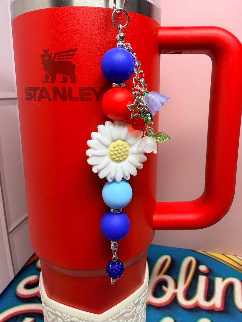 Op de afbeelding: Een rode Stanley-beker met een decoratieve bedel. De bedel heeft blauwe, rode en witte kralen, een witte madelief en kleine bloemaccenten. De beker heeft een handvat en het Stanley-logo.