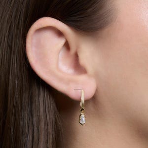 May include: A close-up of a gold hoop earring with a small, dangling, teardrop-shaped pendant. The pendant features a clear, faceted gemstone. The earring is worn on a person's ear.