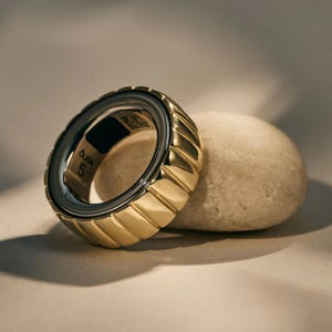 Könnte beinhalten: Ein goldfarbener Ring mit geripptem Design und dunklem Innenband. Der Ring liegt auf einem glatten, hellfarbenen Stein. Der Text "Oura 5" ist auf dem Ring sichtbar.