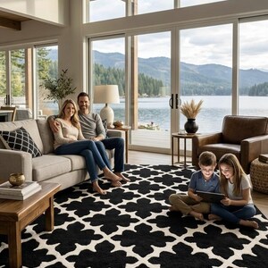 Könnte beinhalten: Wohnzimmer mit einem schwarz-weiß gemusterten Teppich. Eine Familie sitzt auf einem hellgrauen Sofa, und zwei Kinder benutzen ein Tablet. Große Fenster bieten einen Blick auf einen See und Berge.