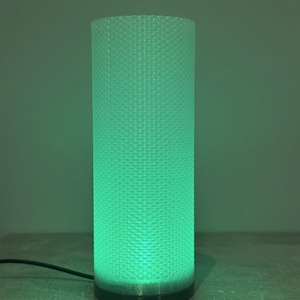 Peut inclure: Une lampe cylindrique à surface texturée, émettant une lueur verte vive. La lampe a une base sombre et un cordon d'alimentation. Le design est moderne et minimaliste.