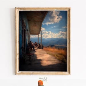 May include: A framed print of a porch scene with people, mountains, and a blue sky. The porch has a wooden floor and a blue wall. The print is in a gold frame. The image is from Moonlit & Laurel Designs.