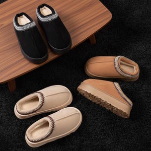 Puede incluir: Una colección de zapatillas cómodas en varios colores. Dos zapatillas negras con ribete blanco están sobre una superficie de madera. Otras zapatillas son de color beige y marrón claro, con un interior suave y afelpado y ribetes decorativos. Las zapatillas están sobre una alfombra oscura.