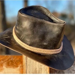 May include: A black leather cowboy hat with a braided tan band. The hat has a wide brim and a slightly distressed appearance, suggesting a vintage or rustic style. The hat is resting on a wooden surface.
