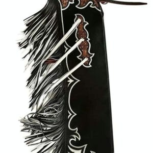 Könnte beinhalten: Schwarze Lederchaps mit weißem und schwarzem Fransenbesatz. Die Chaps haben eine dekorative weiße Zierleiste und braune Lederakzente. Ein Lederriemen erstreckt sich von oben.