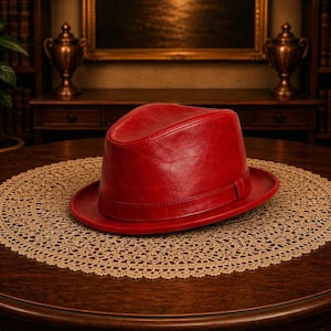 Könnte beinhalten: Ein leuchtend roter Fedora-Hut mit glatter, polierter Oberfläche und einem passenden roten Band, das die Basis der Krone umschließt. Der Hut verfügt über ein klassisches Design mit eingekniffener Krone und einer strukturierten Krempe.