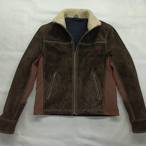 Può includere: Giacca in pelle scamosciata marrone con colletto in montone color crema. La giacca presenta una chiusura con zip frontale, due tasche con zip e dettagli di cucitura marroni. Le maniche hanno un polsino marrone. La giacca ha un pannello laterale marrone e rosa.