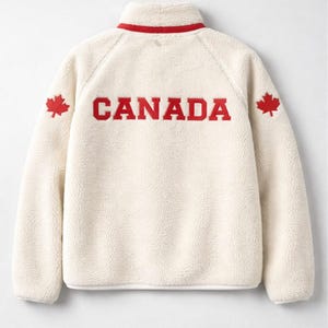 May include: Cream fleece jacket with a red collar and the word "CANADA" in red block letters across the back. Red maple leaf emblems are on each shoulder. The jacket has a high collar.