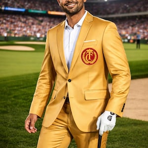 May include: A man in a gold suit and white Titleist cap stands on a golf course. The suit has a red and gold emblem on the chest. He wears a white shirt and glove, holding a golf club.