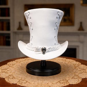 Puede incluir: Un sombrero de copa blanco con ala ancha, con costuras decorativas y una banda trenzada. Un dije de calavera plateada adorna el frente. El sombrero se exhibe sobre un soporte negro, mostrando su diseño único.