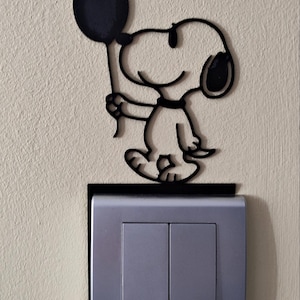 Può includere: Una decorazione da parete nera, piatta e sagomata, che raffigura il personaggio dei cartoni animati Snoopy mentre tiene in mano un palloncino. L'oggetto è montato direttamente sopra una placca rettangolare per interruttore doppio color argento.
